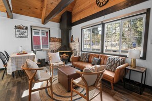 Un salon chaleureux avec une cheminée, des meubles en bois et de grandes fenêtres donnant sur l’extérieur.