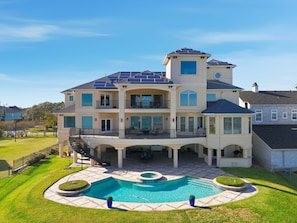 Uma casa grande, de vários andares, com piscina, várias varandas e painéis solares no telhado.