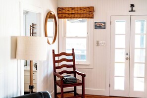 A room with a wooden chair, a lamp, a mirror, a window with a view, and a door with a light switch. - 2BR Folly Cottage 1-block from the beach! (Folly Beach)