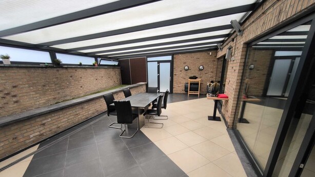 Un patio al aire libre con una mesa larga y sillas, una pared de ladrillo y un ventanal de vidrio que da a otra área.