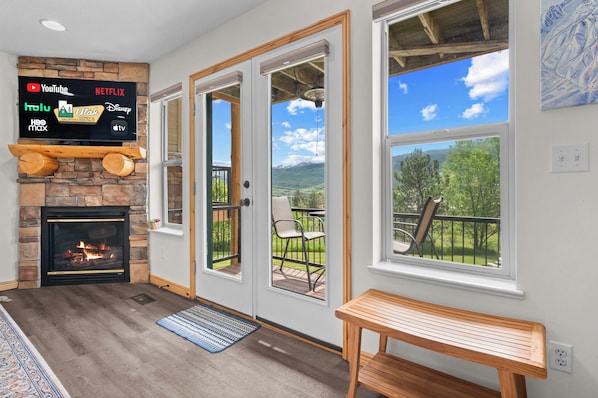 A room with a stone fireplace, a flat-screen TV displaying streaming services, a balcony with outdoor seating, and a large window offering a mountain view.
