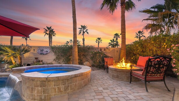 Un jacuzzi con caída de agua, rodeado de palmeras y un patio con sillas y un pozo de fuego.