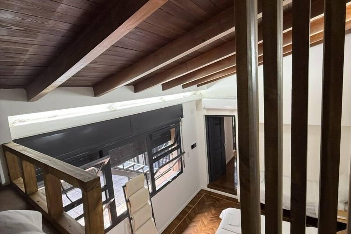 A loft-style bedroom with a high ceiling, wooden beams, a herringbone-patterned floor, and a view of the street through a large window.