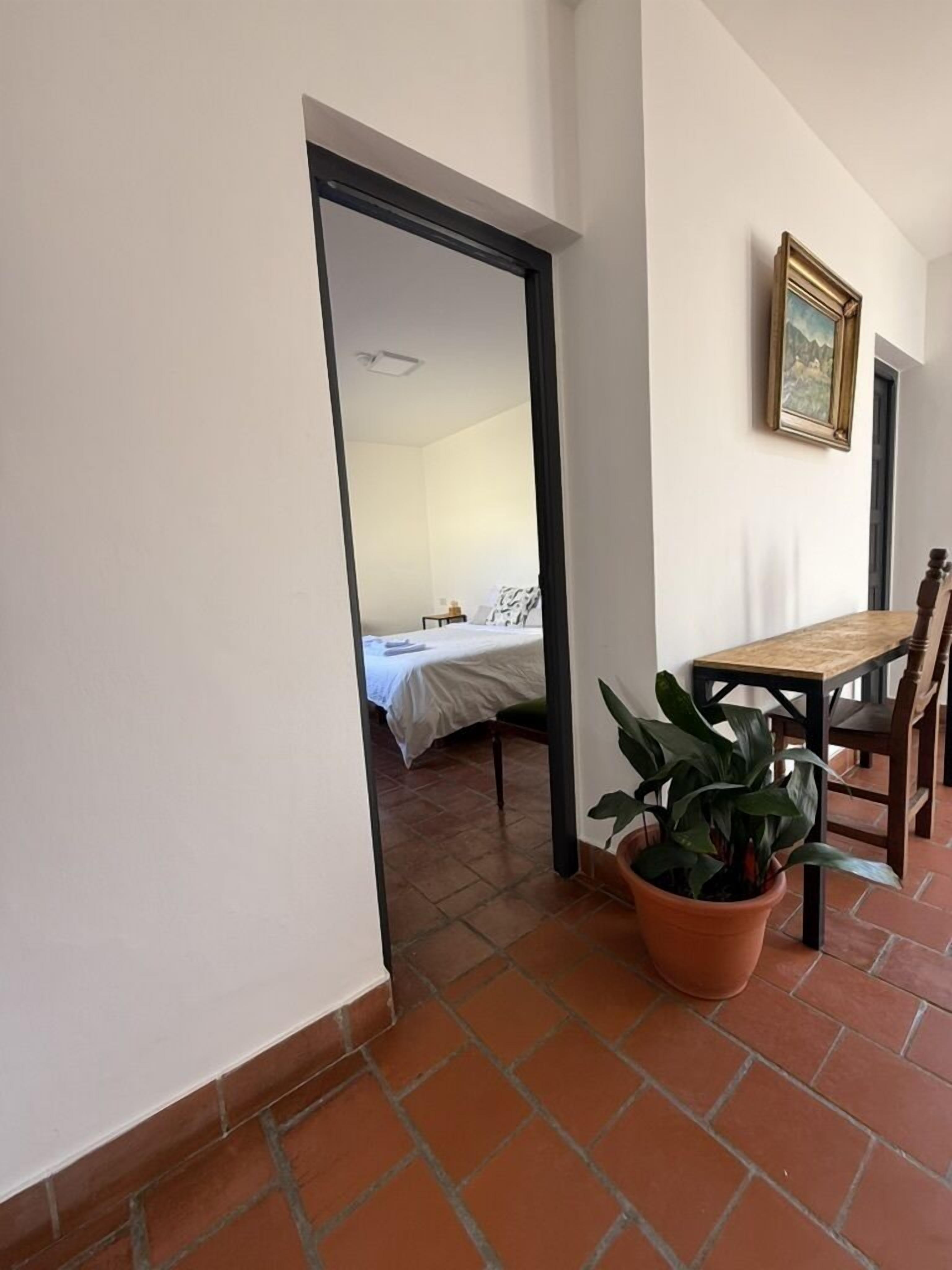 A corridor leading to a bedroom with a bed, a small table, and a chair. There is a potted plant in the corridor.