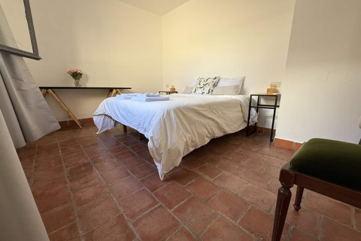 A bedroom with a bed, a bedside table, a chair, and a shelf with a vase.