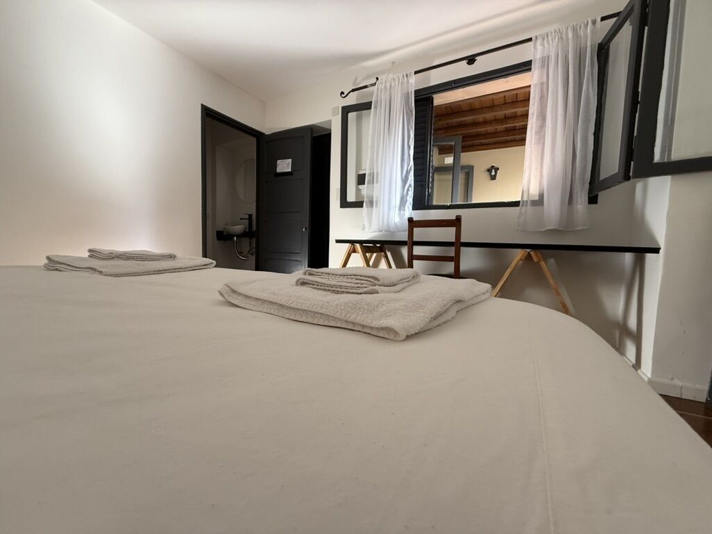 A bedroom with a large bed, a desk, and a chair. There is a window with curtains, a door, and a mirror on the wall.