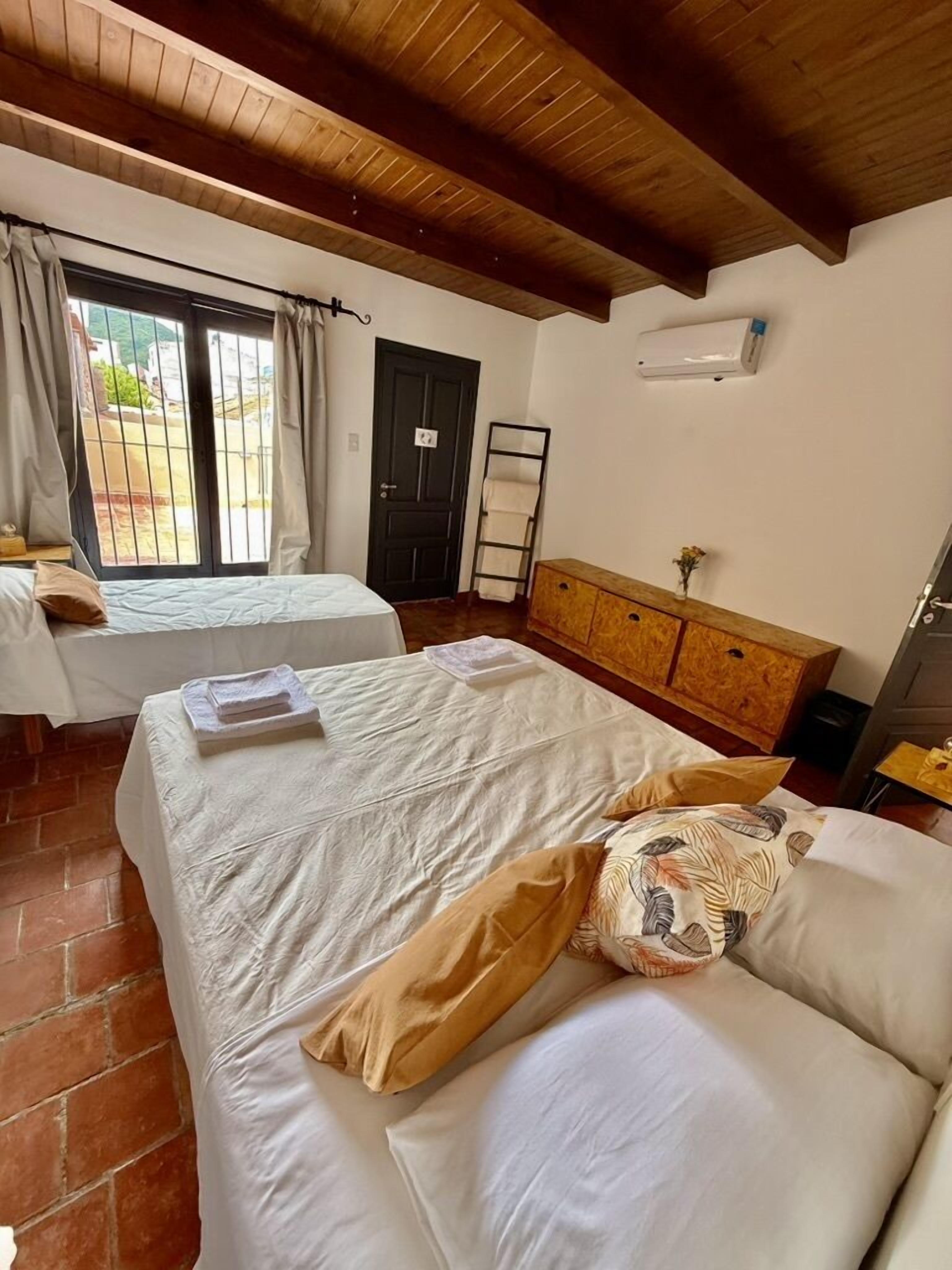 A bedroom with two beds, a wooden ceiling, a window with curtains, a door, a wooden dresser, and a wall-mounted air conditioner.
