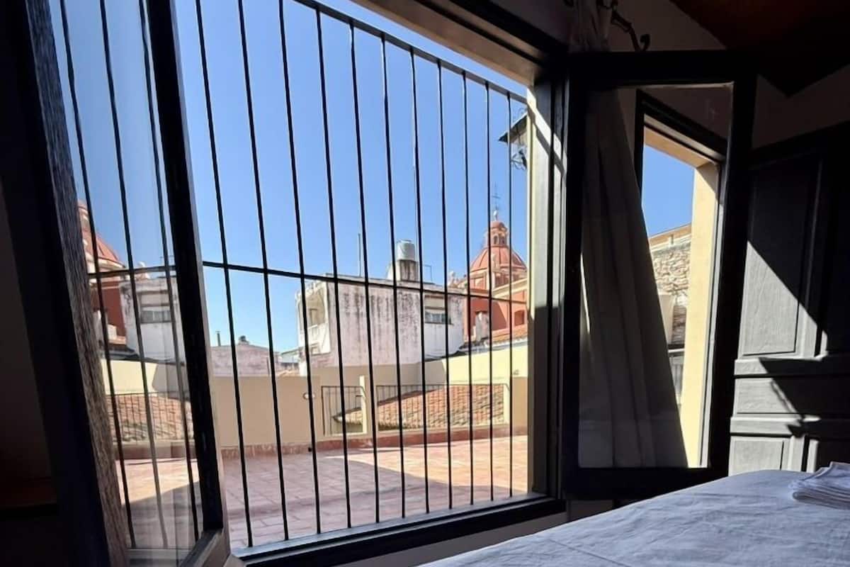 A room with a bed, a window with metal bars, and a view of buildings outside.