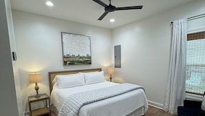 Un dormitorio con una cama, ventilador de techo, mesita de noche con lámpara, un cuadro en la pared y ventana con cortinas.