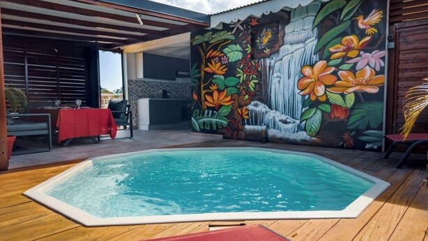 A swimming pool with a mural of tropical flowers and waterfalls, surrounded by wooden decking and a covered seating area. - Le Havre de Paix (Le Moule)