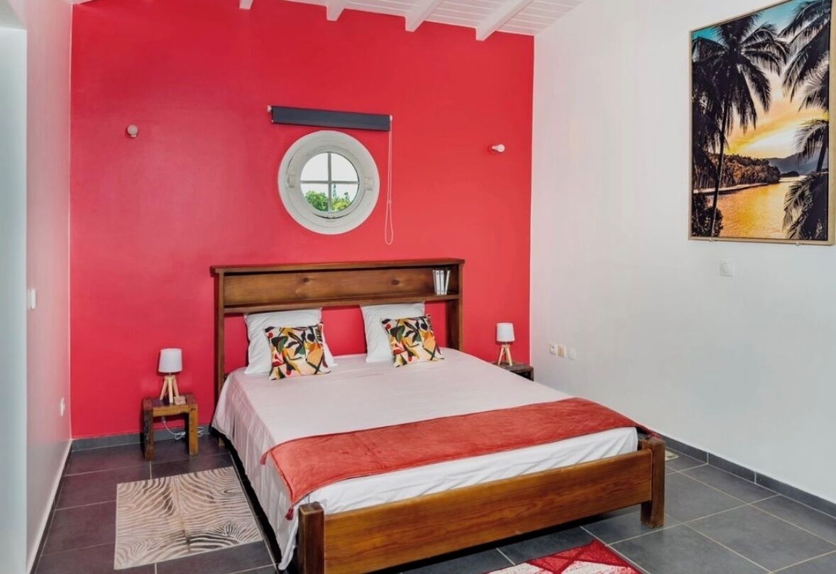 A bedroom with a red wall, a round window, a bed with a wooden headboard, two bedside tables with lamps, and a painting of a beach scene.