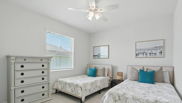 Une chambre avec deux lits, une commode, un ventilateur de plafond et une fenêtre avec des stores.