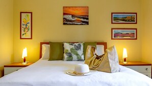 Ein ordentlich bezogenes Bett mit weißer Bettwäsche, ein weißer Hut und zwei Nachttische mit Lampen. An der Wand hängen drei gerahmte Bilder.