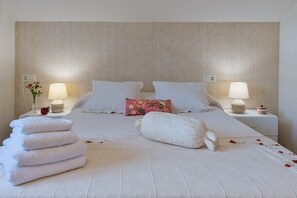 Cama bem arrumada com lençóis brancos, um travesseiro decorativo e uma pilha de toalhas. Há duas luminárias de cabeceira e um vaso com flores na mesinha de cabeceira.