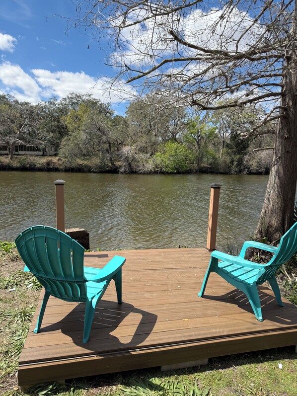 A wooden deck with two turquoise chairs facing a calm river, surrounded by trees and a clear sky. - Cozy Waterfront Cabin Minutes from the Beach (Palacios)