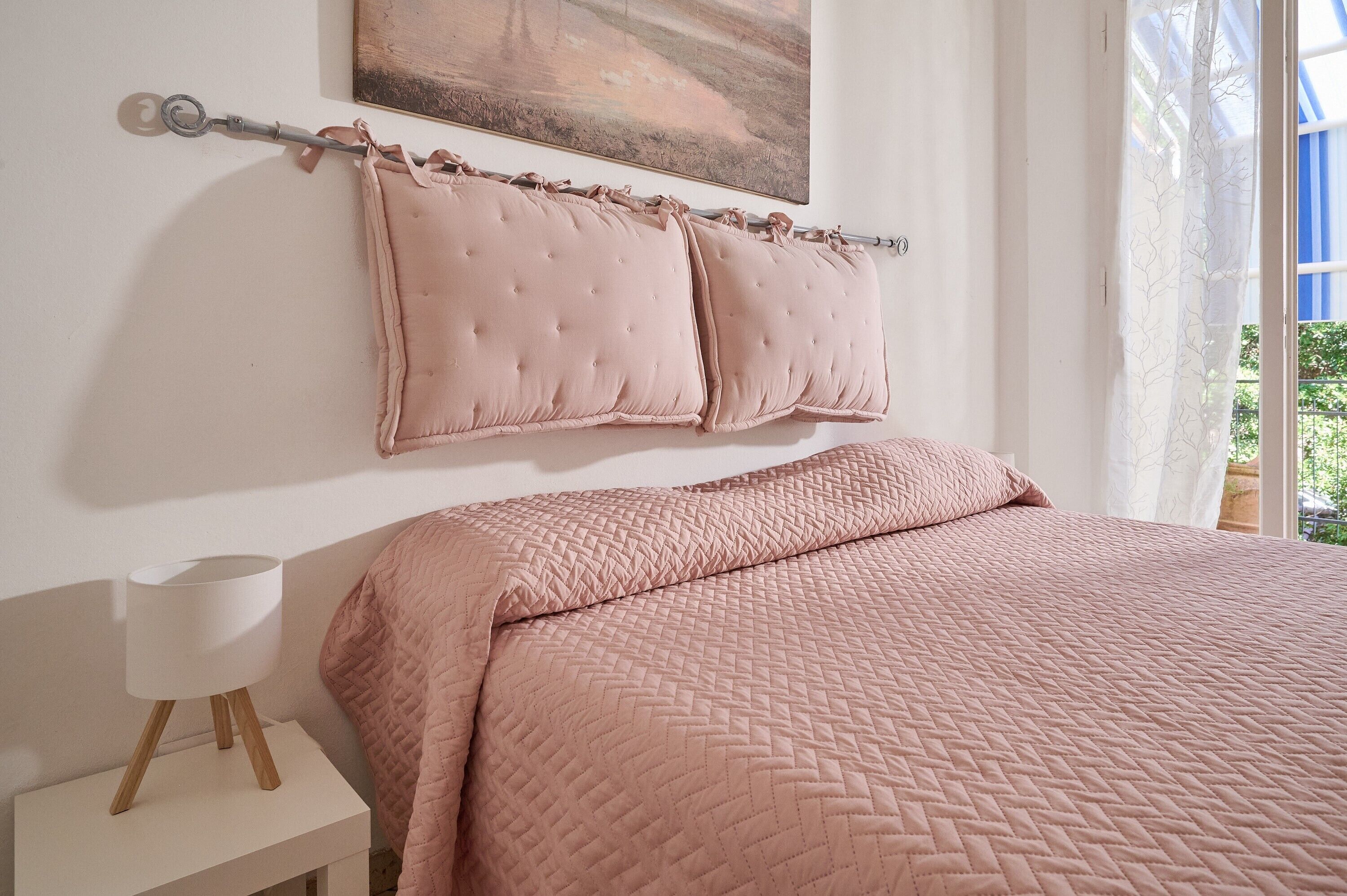 A bed with a pink quilted bedspread, a white bedside table with a lamp, and a painting on the wall.