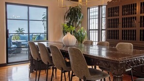 A dining area with a large wooden table, six chairs, a chandelier, a potted plant, and a view through large windows. - Epic Resort Villa: Pool, Spa, Firepit, Ocean View, Games (San Diego)