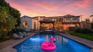 A swimming pool with a pink flamingo float, a covered patio area, and a house with a tiled roof. - Epic Resort Villa: Pool, Spa, Firepit, Ocean View, Games (San Diego)