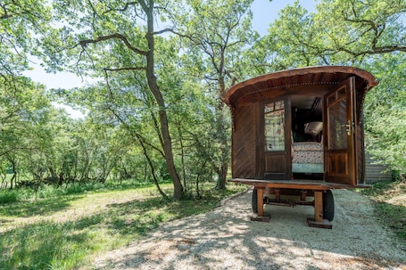 A wooden cabin with an open door, a bed inside, and a gravel path leading to it.. Cottage 'La Roulotte Authentique' with Private Terrace, Private Garden and Wi-Fi