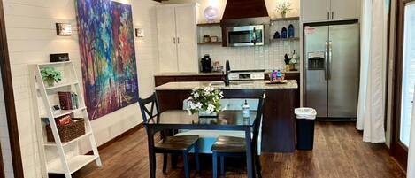 Una cocina moderna con piso de madera, muebles de cocina blancos, una mesa de comedor con sillas, un cuadro en la pared y un refrigerador.