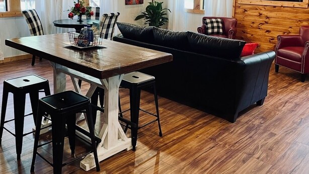 Ein gemütliches Blockhaus-Interieur mit Holzbalkendecke, einem Essbereich mit Tisch und Stühlen, einem schwarzen Ledersofa und einem an der Wand montierten Fernseher.