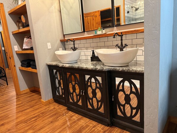 Un baño con dos lavamanos tipo recipiente, estantes de madera, un espejo y suelo de madera.