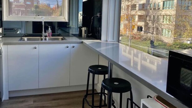 A modern kitchen with a large window, a sink, a clock, and two bar stools.