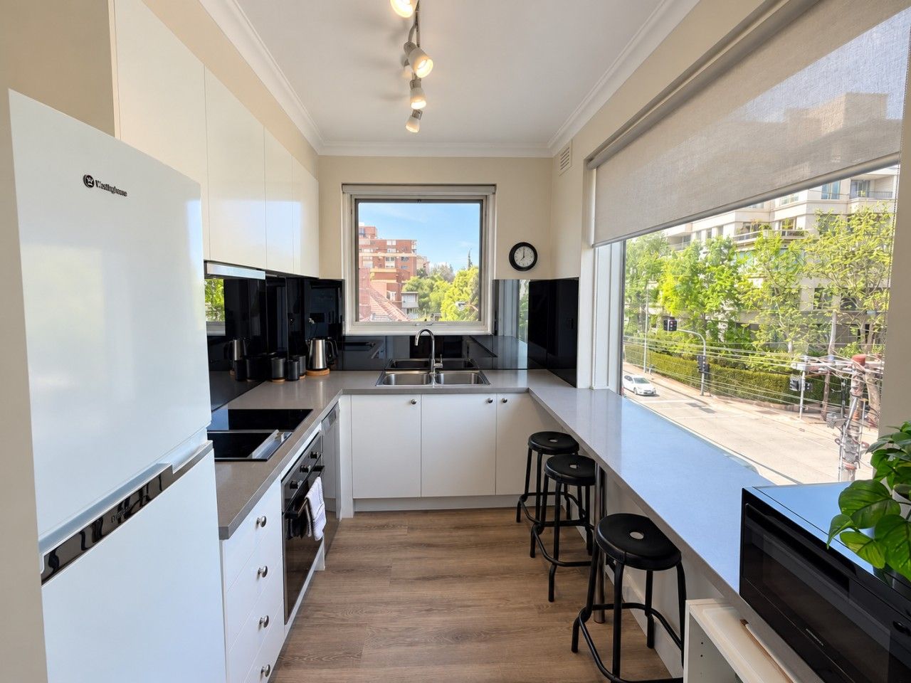 Une cuisine moderne avec des armoires blanches, un comptoir noir, une cuisinière intégrée et une fenêtre donnant sur des immeubles et des arbres.