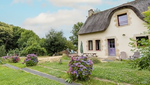 Ein rustikales Haus mit Strohdach, einer kleinen Veranda und einem Garten mit blühenden Büschen.