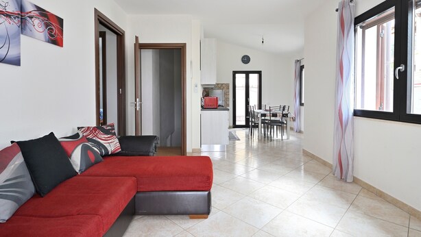 Uma sala de estar com um sofá vermelho, um espaço de jantar com mesa e cadeiras, e uma cozinha ao fundo.
