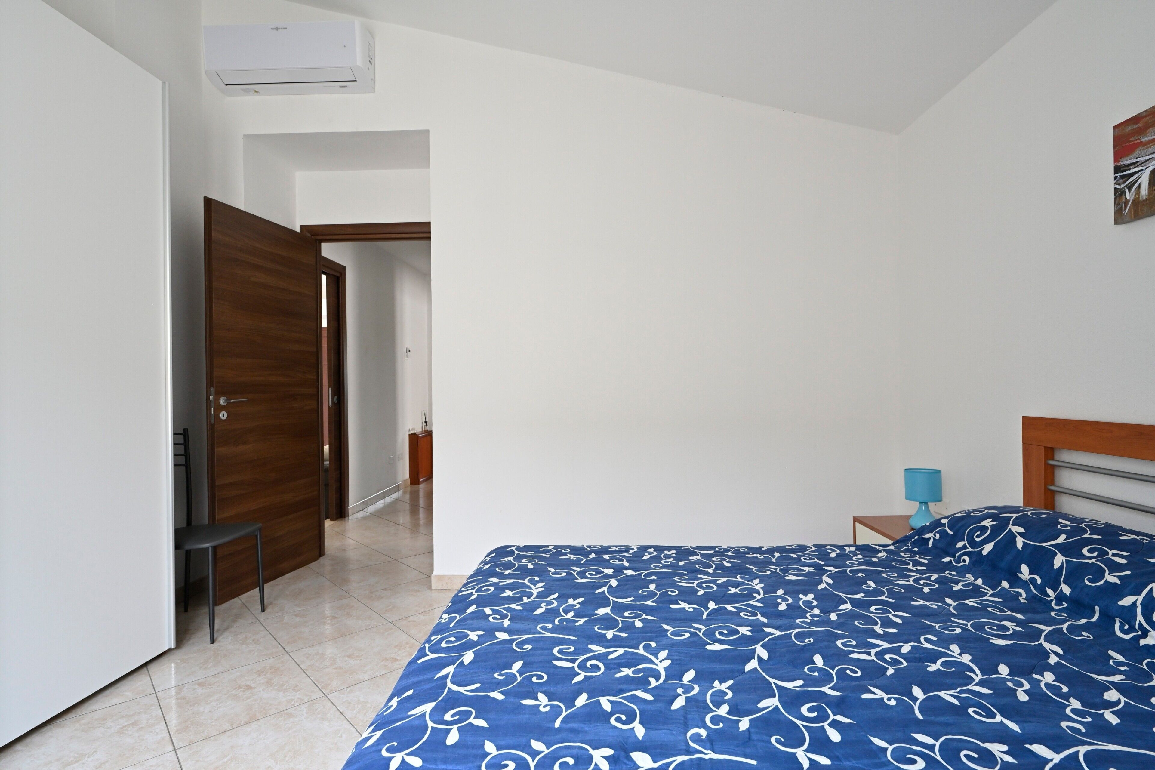 Ein Schlafzimmer mit Bett, Stuhl, Holztür, weißer Wand und einem kleinen Tisch mit Lampe.