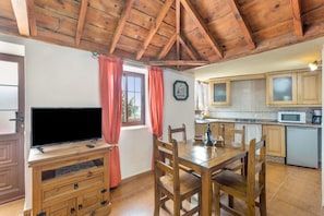A dining area with a wooden table and chairs, a television on a wooden stand, and a kitchen with cabinets and a microwave.