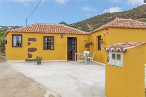 A yellow building with a tiled roof, a small patio with a table and chairs, and a potted plant.