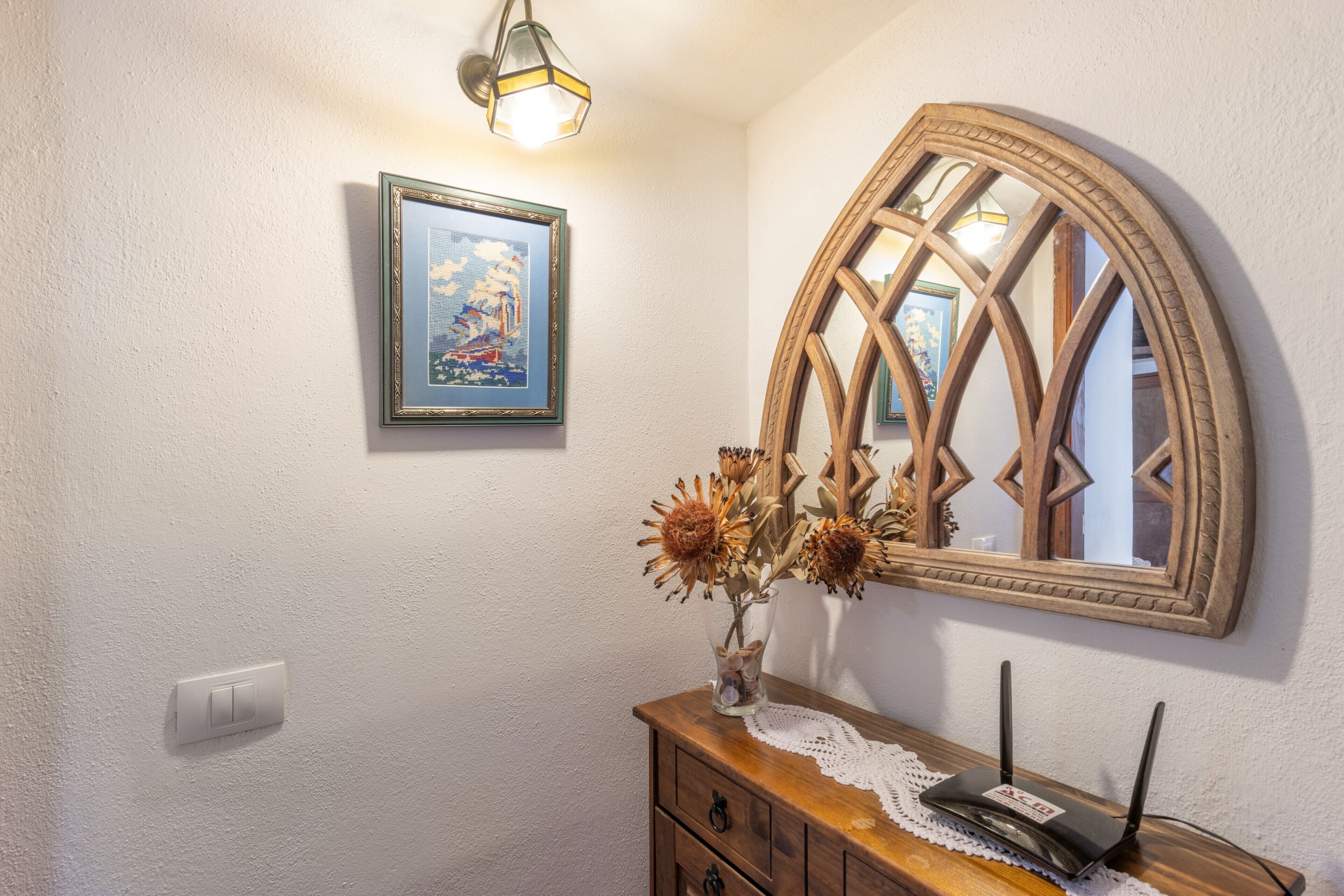 Una habitación con un aparador de madera, un cuadro enmarco, un jarrón con flores secas y un espejo con diseño geométrico.