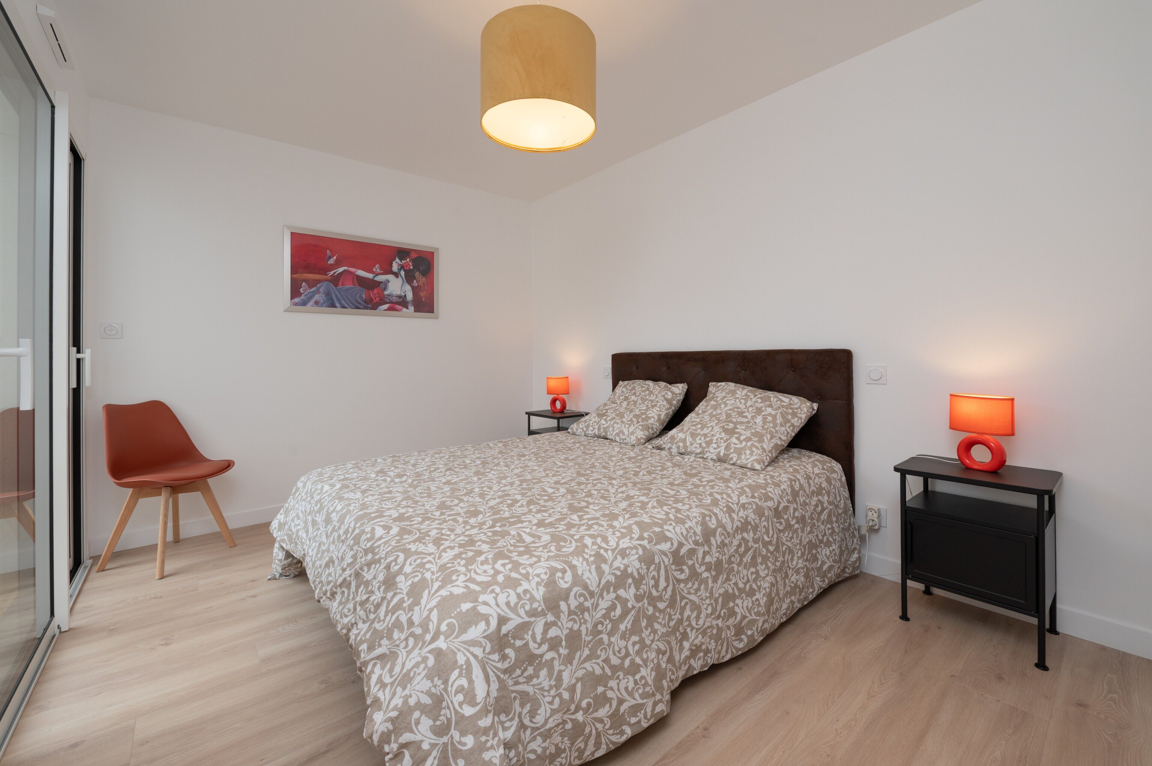 A bedroom with a bed, two bedside tables with lamps, a chair, a painting on the wall, and a wooden floor.