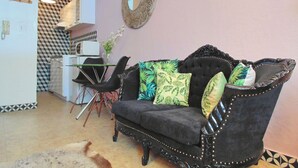 Ein Wohnzimmer mit einem dunkel gepolsterten Sofa, einem Esstisch mit Stühlen, einer Küche im Hintergrund und einem gemusterten Boden.