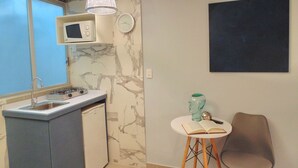 Eine kleine Küche mit Spüle, Mikrowelle und kleinem Kühlschrank. Es gibt einen Essbereich mit einem Stuhl und einem Tisch sowie eine an der Wand befestigte Uhr.