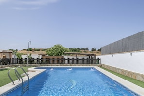 A rectangular swimming pool with a wooden deck and metal railings, surrounded by a white wall and a fence.