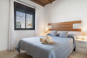 A neatly made bed with a blue quilt, a wooden headboard, a nightstand with a lamp, and a window with a view of buildings.