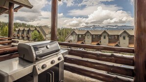 A wooden deck with a grill, overlooking a row of cabins with a mountainous backdrop.