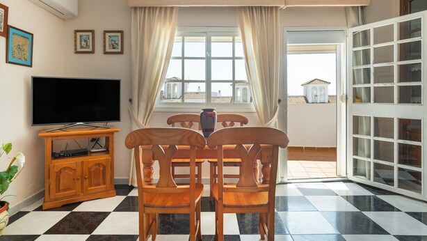 Una habitación con televisión de pantalla plana, sillas de madera, una mesa de comedor y un ventanal con cortinas.