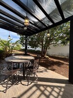 An outdoor patio with a round table and chairs, under a pergola with hanging lights.