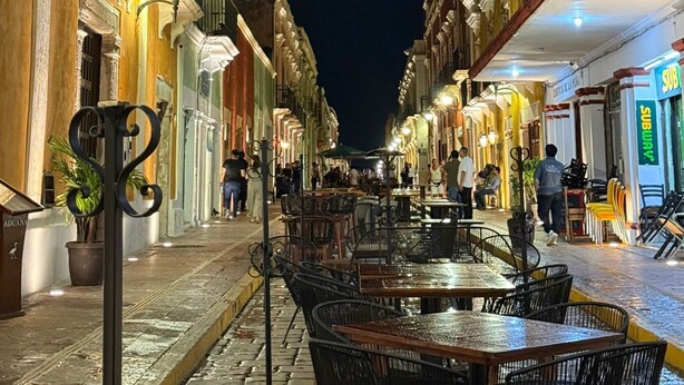 Una calle nocturna con mesas al aire libre, edificios con balcones y un restaurante Subway.