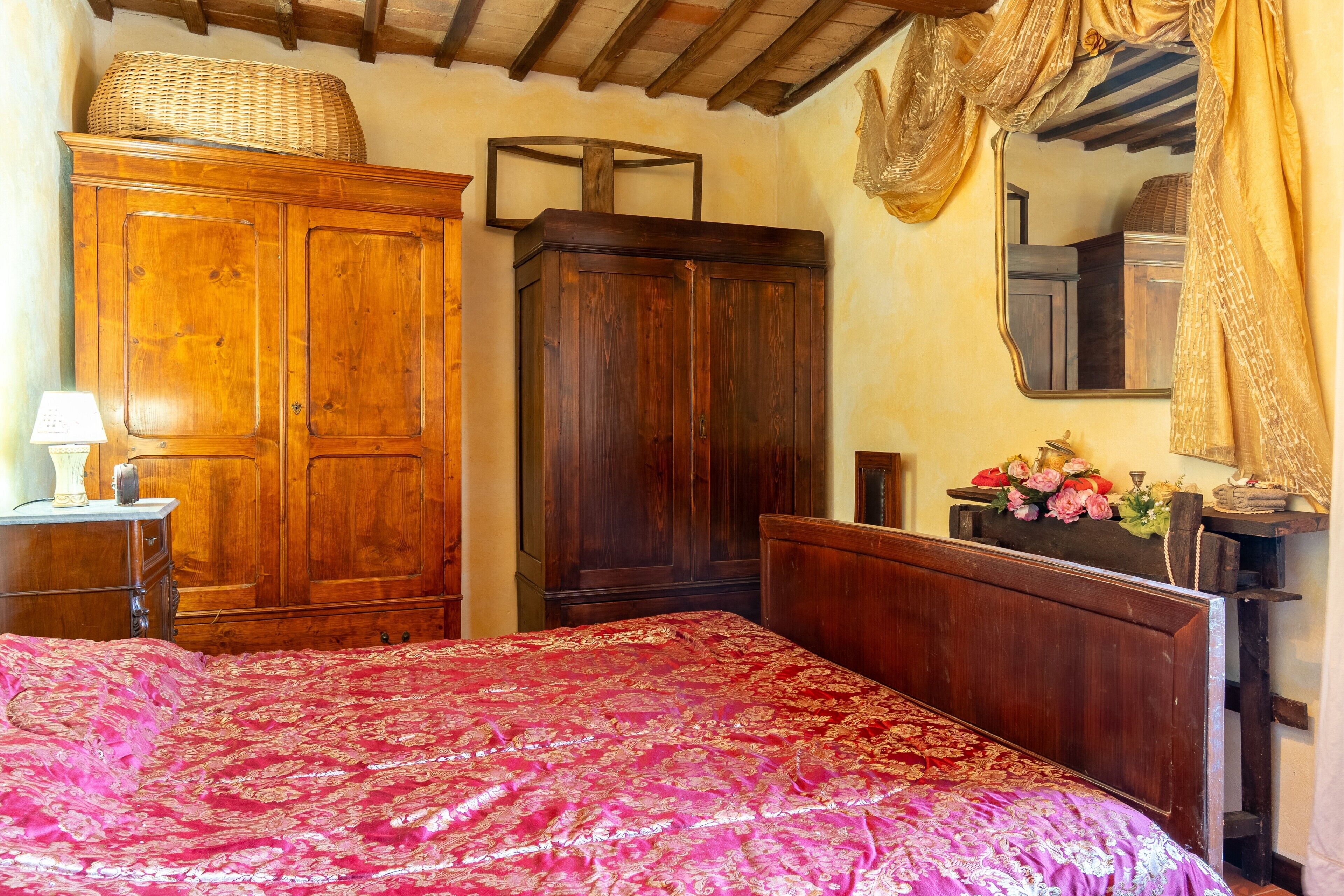 A bedroom with a large wooden bed, a wardrobe, a mirror, and a small table with a lamp.