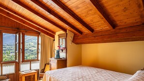Un dormitorio con techo de madera, cama, mesita de noche, ventana con cortinas y vistas a un paisaje montañoso.
