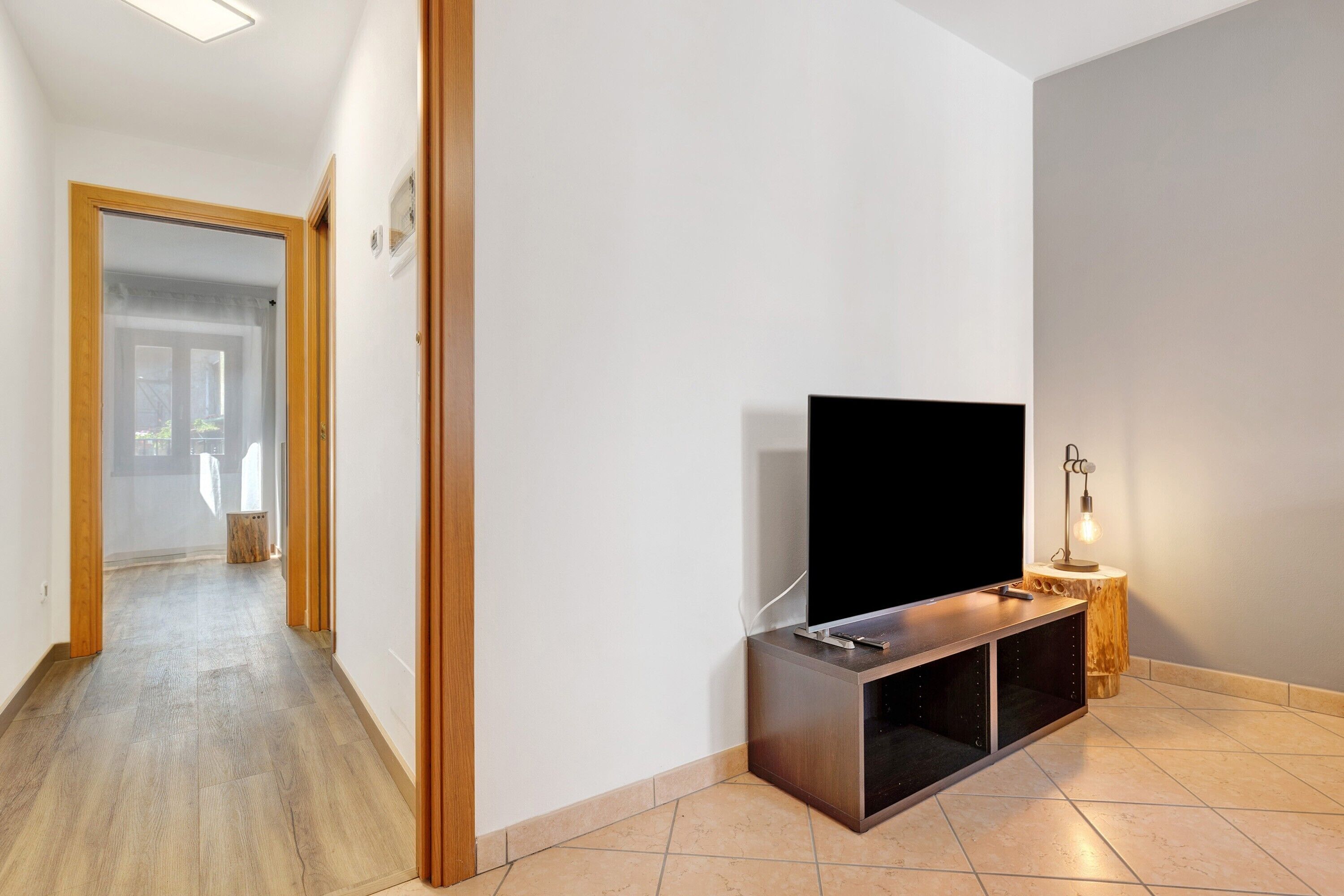 Ein Flur, der zu einem Raum führt, in dem sich ein Flachbildfernseher auf einem Holzboden, ein kleiner Holzschemel mit einer Lampe und eine Tür zu einem anderen Raum befinden.