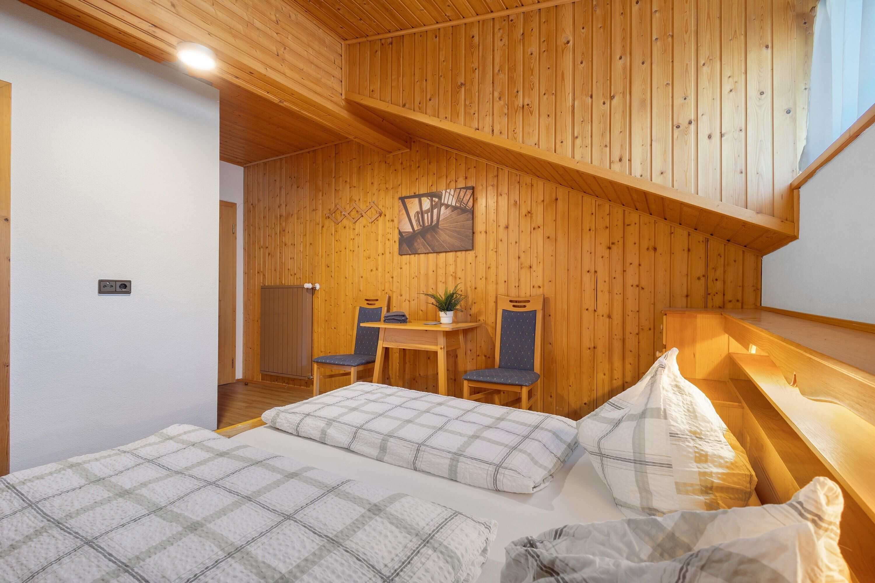 A bedroom with a wooden ceiling, a bed with a plaid duvet, a desk with two chairs, and a small plant.