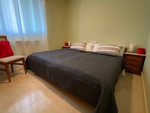 A bedroom with a bed, a chair, a nightstand, and a bedside table.