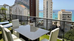 Un balcón con mesa y sillas, con vistas a una ciudad costera con edificios y una montaña.