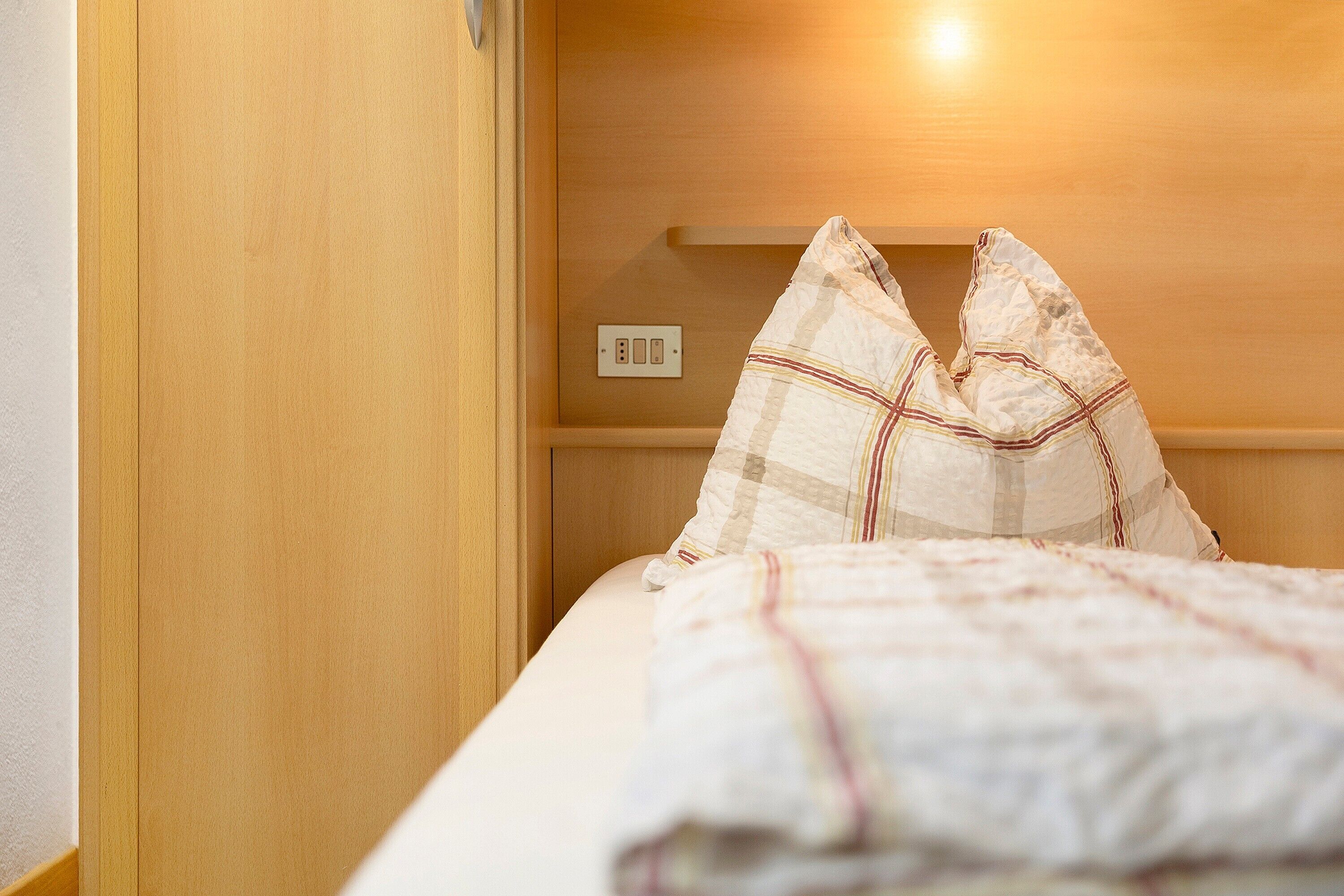 A bed with a checkered blanket and pillows, a wooden wardrobe, and a wall socket.